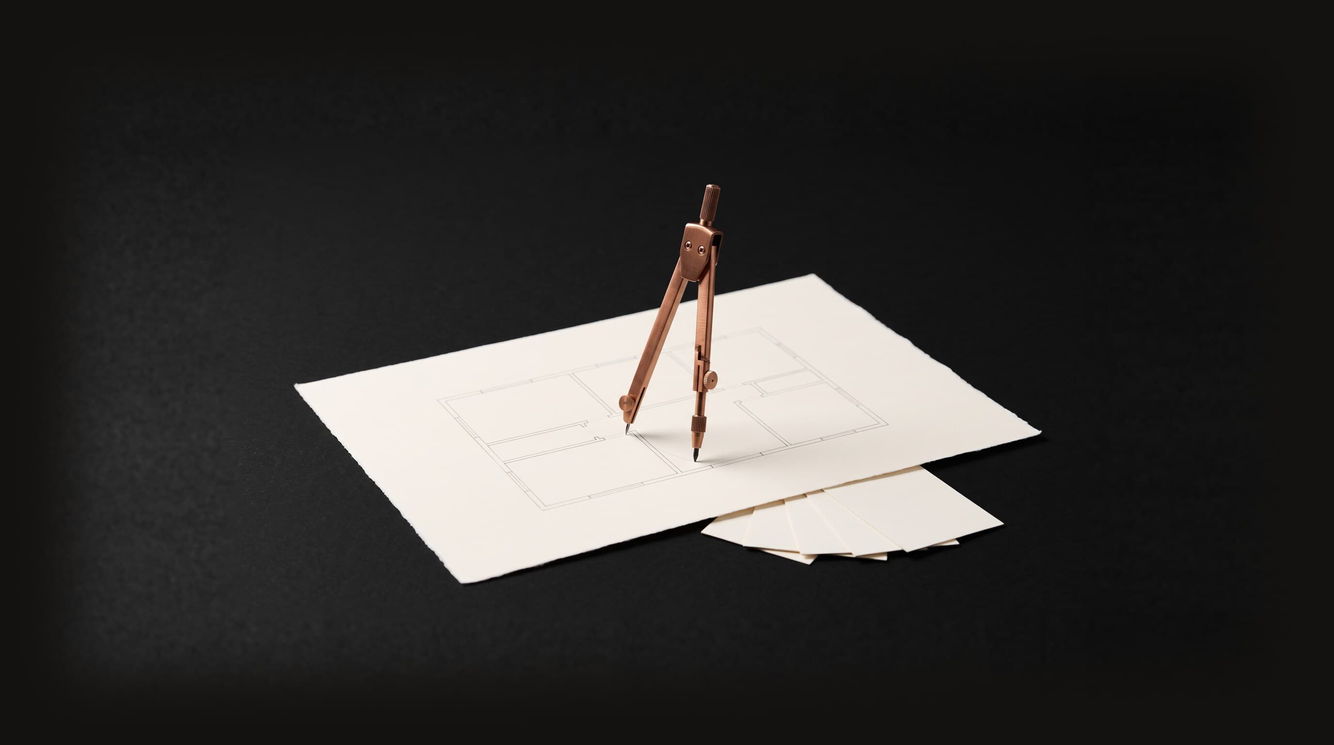 A copper drafting compass standing on a large cream sheet that carries a faint hand-drawn floor plan, with five small blank cream cards tucked under one edge, representing a plan drawn before anyone starts.