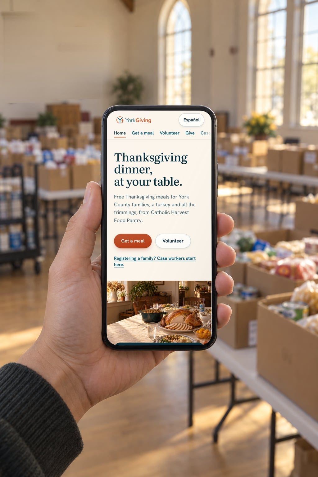 A hand holding a phone in a community hall, showing the yorkgiving.com home page: its headline, buttons to get a meal and to volunteer, and a link for case workers.