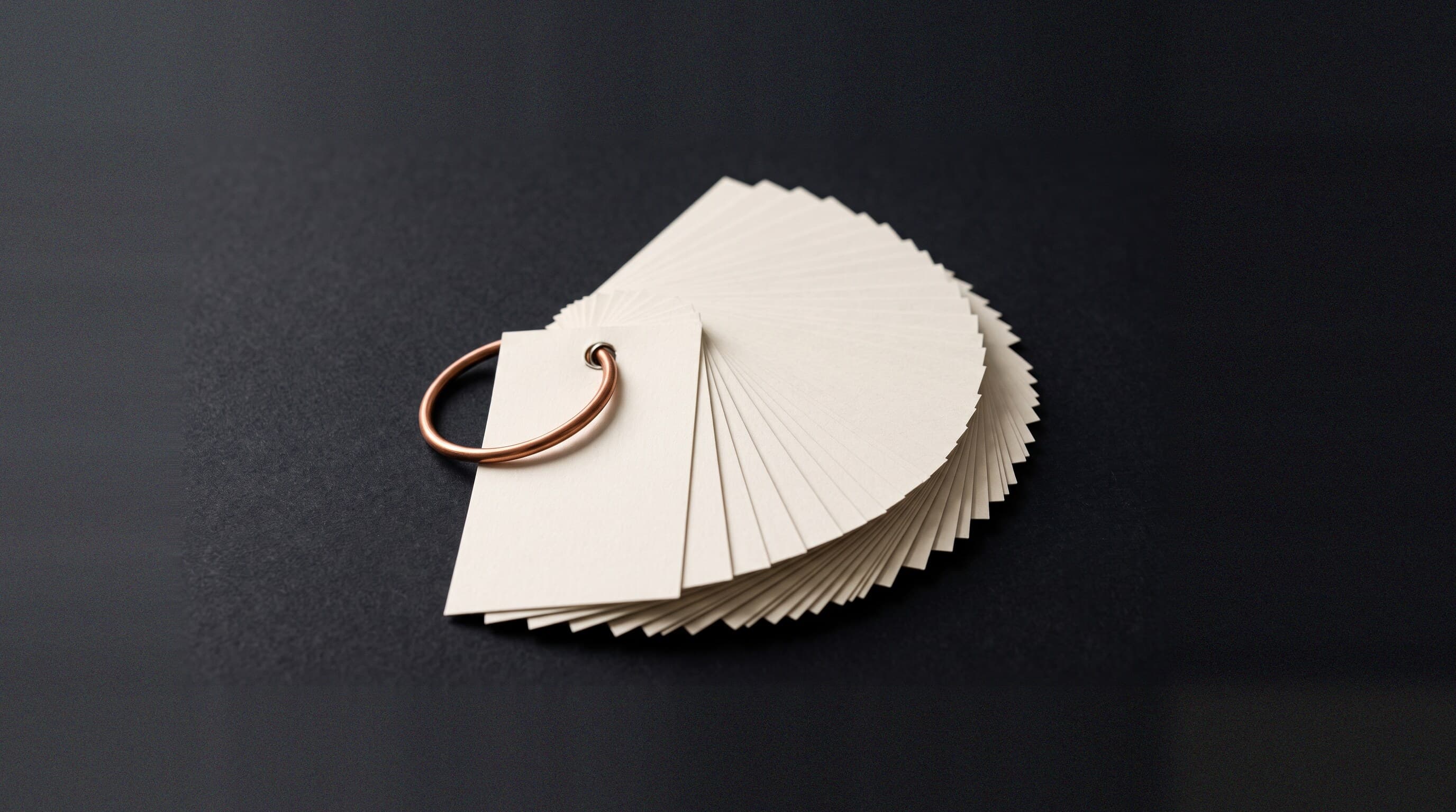 A copper ring threaded through a fanned deck of blank cream index cards on a dark charcoal field, representing many separate terms bound into one working reference.