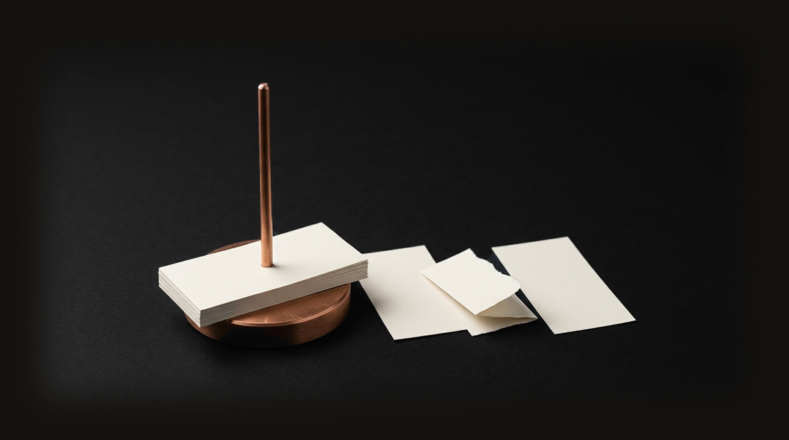 A copper desk paper spike holding a stack of blank cream slips, with three more blank slips lying beside it on a dark field, one of them smaller, folded, and torn along one edge, representing an AI that takes in everything that reaches it, including the note somebody else slipped in.